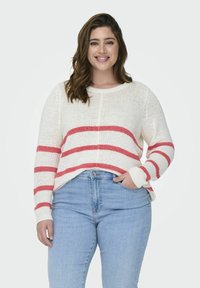 Maglione bianco a maglia con strisce orizzontali rosse, scollatura tonda e maniche lunghe, abbinato a jeans azzurri chiari. La texture appare morbida e accogliente.