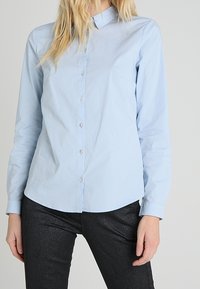 Chemise bleu clair à manches longues, coupe ajustée, avec un col et une texture lisse, accompagnée d'un pantalon sombre et brillant.