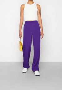 Fialové široké kalhoty s elastickým pasem, kombinované s bílým topem bez rukávů a malou žlutou kabelkou. Bílé tenisky dokončují celý outfit.