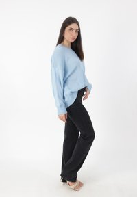 Helle blaue, übergroße Strickjacke mit gerippten Bündchen, kombiniert mit schwarzen geraden Jeans und beigen Riemchen-Heels. Weißer Hintergrund.