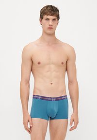 Männlicher Model mit kurzen braunen Haaren, der türkisfarbene Boxershorts mit lila Bund und "Sloggi"-Logo trägt und vor einem schlichten weißen Hintergrund steht.