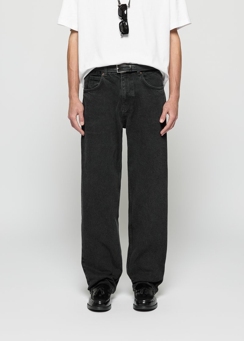 Person, die lockere schwarze Jeans, einen schwarzen Gürtel, ein weißes kurzärmliges Hemd, schwarze Quasten-Slipper und eine Sonnenbrille, die am Kragen des Hemdes hängt, trägt.
