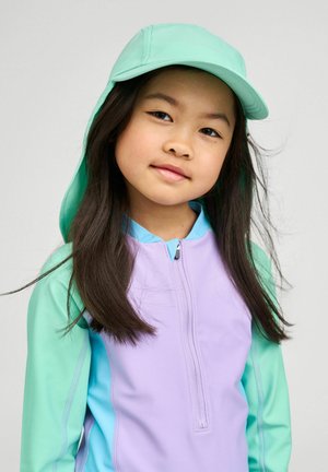 Młoda dziewczyna z długimi ciemnymi włosami, ubrana w pastelową fioletową i miętowo zieloną kurtkę oraz pasujący miętowo zielony kapelusz przeciwsłoneczny, zwrócona przodem.