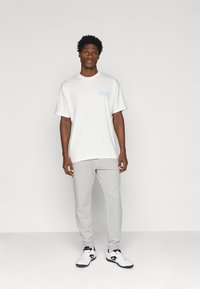 Camiseta blanca con "City" en un script azul claro, combinada con pantalones de chándal grises. Zapatillas en blanco y negro con acentos dorados completan el conjunto.