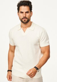 Cremefarbener Polo-Shirt mit kurzen Ärmeln und strukturiertem Muster, V-Ausschnitt und geripptem Kragen. Als Accessoires sind eine schwarze Uhr und ein Armband vorhanden.