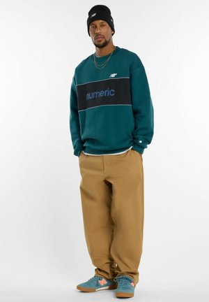 Mann trägt einen türkisfarbenen "Numeric"-Pullover, beige Hose, türkis-orangefarbene Turnschuhe und eine schwarze Mütze, steht mit den Händen in den Taschen vor weißem Hintergrund.