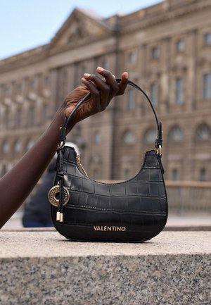 Main à la peau foncée tenant un sac à main Valentino noir avec des accents dorés devant un bâtiment historique flou en arrière-plan.