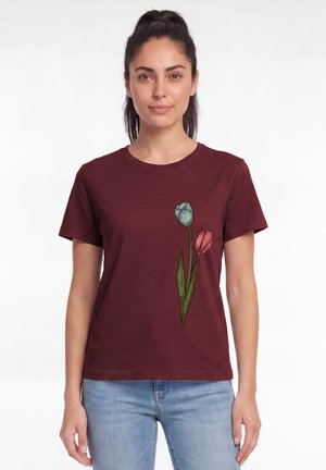 Frau mit dunklen Haaren im Pferdeschwanz, die ein kastanienbraunes T-Shirt mit blauen und pinken Tulpenmotiven und hellblaue Jeans vor weißem Hintergrund trägt.