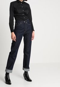 Černá košile na knoflíky nošená s tmavými džínami s vysokým pasem, zakončenými ohrnutými lemy, a lesklými černými kotníkovými botami.