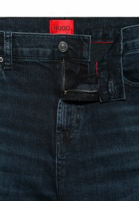 Pantalones vaqueros de mezclilla azul oscuro con bragueta de cremallera, que cuentan con un bolsillo delantero y una etiqueta de marca roja con la inscripción "HUGO". Textura suave y diseño clásico.
