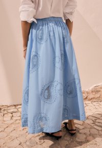 Gonna lunga di cotone azzurro chiaro con motivi paisley ricamati, vita elasticizzata e silhouette fluida. Indossata con sandali neri con tacco.