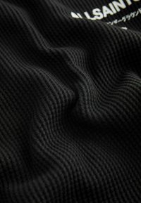 Čierna textúrovaná tkanina s vrúbkovaným vzorom, obsahujúca viditeľné záhyby a potlačenú etiketu s bielym textom. Mäkký, tkaný materiál.