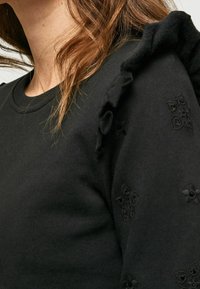 Sudadera negra con detalle de volantes en el hombro y sutil bordado floral en la manga. Tela suave y texturizada con escote redondo.