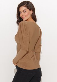Brązowa bluzka z długim rękawem i bufkami na ramionach, z okrągłym dekoltem oraz małym złotym guzikiem z tyłu. Gładki, elastyczny materiał.