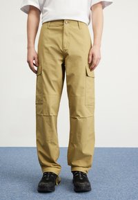 Beige cargo bukser lavet af struktureret stof, med sidelommer, en knaplukning og en løs pasform, parret med sorte sko.