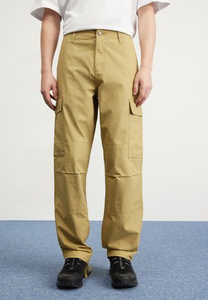 Pantalones cargo beige hechos de tela texturizada, con bolsillos laterales, cierre de botón y un corte holgado, combinados con zapatos negros.