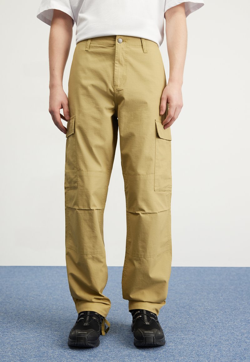 Beige cargo bukser lavet af struktureret stof, med sidelommer, en knaplukning og en løs pasform, parret med sorte sko.
