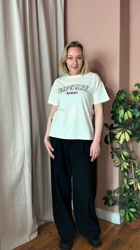 Νεαρή γυναίκα που φοράει λευκό μπλουζάκι με το λογότυπο "RIP CURL HAWAII" και μαύρο φαρδύ παντελόνι, στέκεται μέσα σε εσωτερικό χώρο κοντά σε μπεζ κουρτίνες και γλάστρα με φυτό.