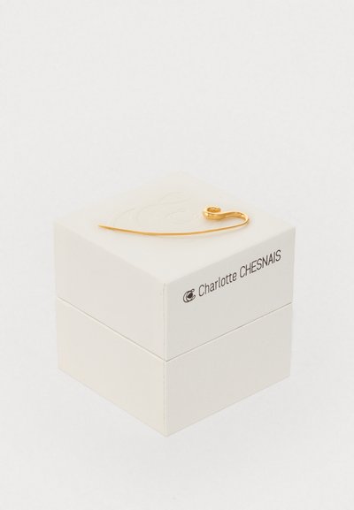 Złota biżuteria w kształcie zakrzywionej szpilki, leżąca na teksturowanym białym pudełku z subtelnym wytłoczonym logo. Pudełko ma gładkie wykończenie.