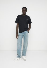 Μαύρο κοντομάνικο μπλουζάκι με λογότυπο στο στήθος, ανοιχτό μπλε τζιν με ίσια μπατζάκια και λευκά αθλητικά παπούτσια με γκρι ανοιχτές λεπτομέρειες, σε λευκό φόντο.