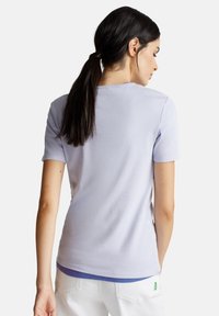 Mujer con cabello oscuro en una coleta, vistiendo una camiseta de mangas cortas de color púrpura claro y pantalones blancos, de espaldas sobre un fondo blanco.
