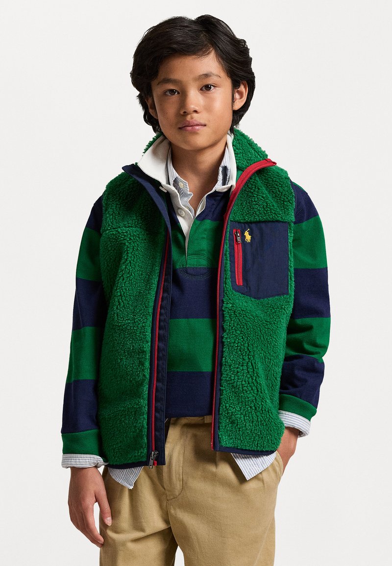 Πράσινη και ναυτική ριγέ φλις ζακέτα με φερμουάρ, κόκκινες πινελιές και τσέπη, φορεμένη πάνω από ένα λευκό πουκάμισο με γιακά και μπεζ παντελόνι.