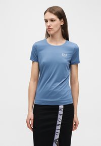 Γυναίκα που φοράει εφαρμοστή μπλε μπλούζα EA7 και μαύρη φούστα με κάθετο λευκό κείμενο Emporio, στέκεται μπροστά σε απλό φωτεινό φόντο.