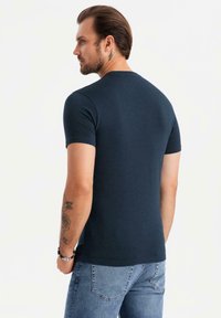 Námořnická modrá přiléhavá tričko s krátkým rukávem, vyrobené z měkké bavlny. Má kulatý výstřih a viditelné tetování na dolní části paže.