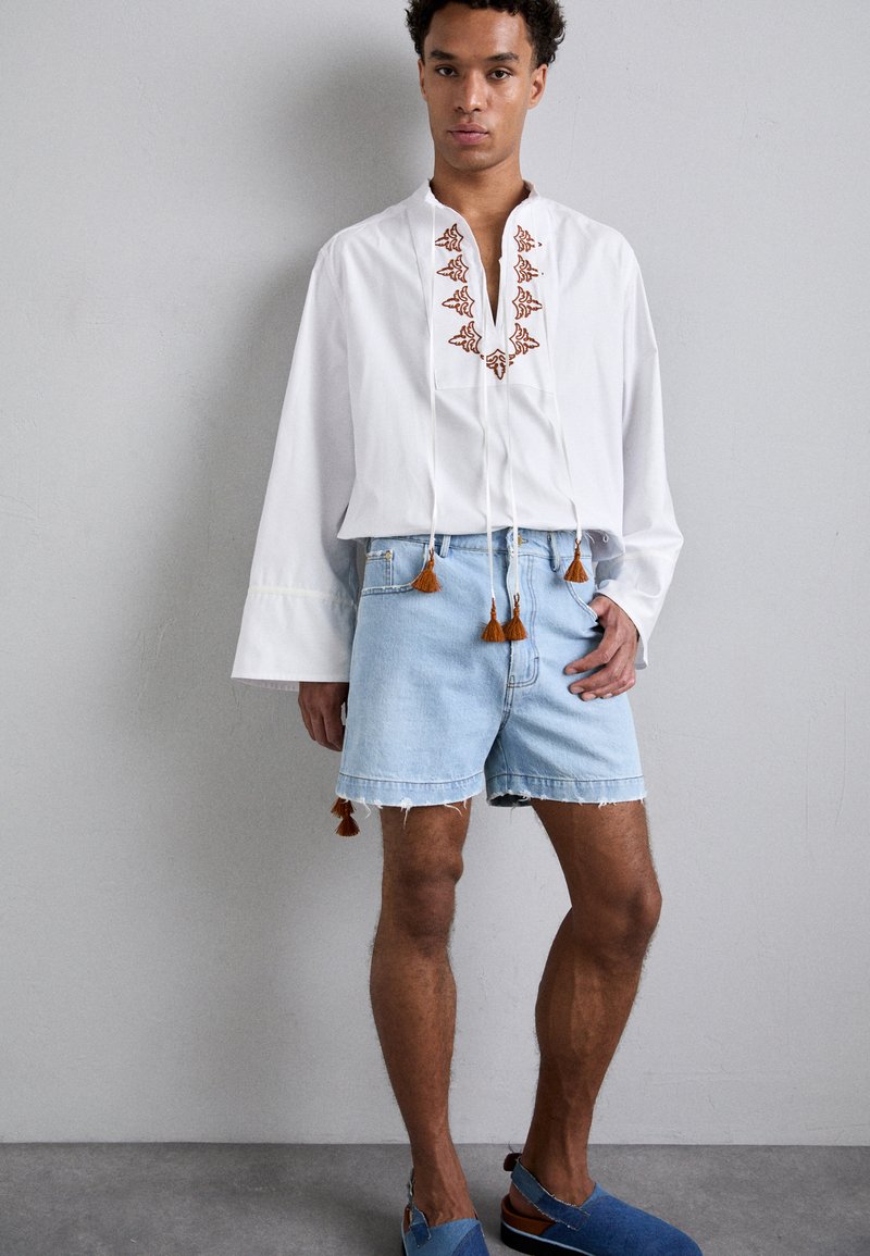 Camisetta bianca a maniche lunghe con ricami marroni e frange, abbinata a pantaloncini di jeans azzurri chiari con bordi sfilacciati e dettagli marroni.