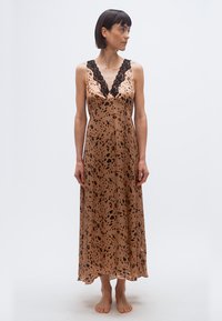 Γυναίκα που στέκεται ξυπόλυτη, φορώντας ένα αμάνικο ταμπά φόρεμα με λεοπάρ σχέδιο και μαύρη δαντέλα στο τελείωμα, κοιτάζοντας προς τα δεξιά της, μπροστά σε απλό λευκό φόντο.