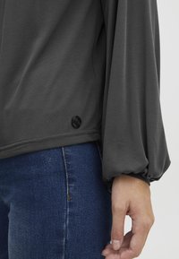 Grå langermet topp med avslappet passform, med en samlet mansjett og en svart knapp detalj nær sømmen, kombinert med mørke denimjeans.
