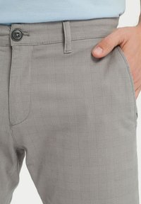 Szare spodnie w kratę z teksturowanej tkaniny, z zapięciem na guzik i kieszenią z przodu. Wzór składa się z kwadratów układających się w siatkę.