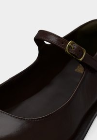 Σκούρο καφέ δερμάτινο παπούτσι Mary Jane με χρυσή πόρπη λουριού πάνω από το πάνω μέρος του ποδιού και στρογγυλεμένη μυτερή μύτη, ορατό σε ανοιχτό φόντο.