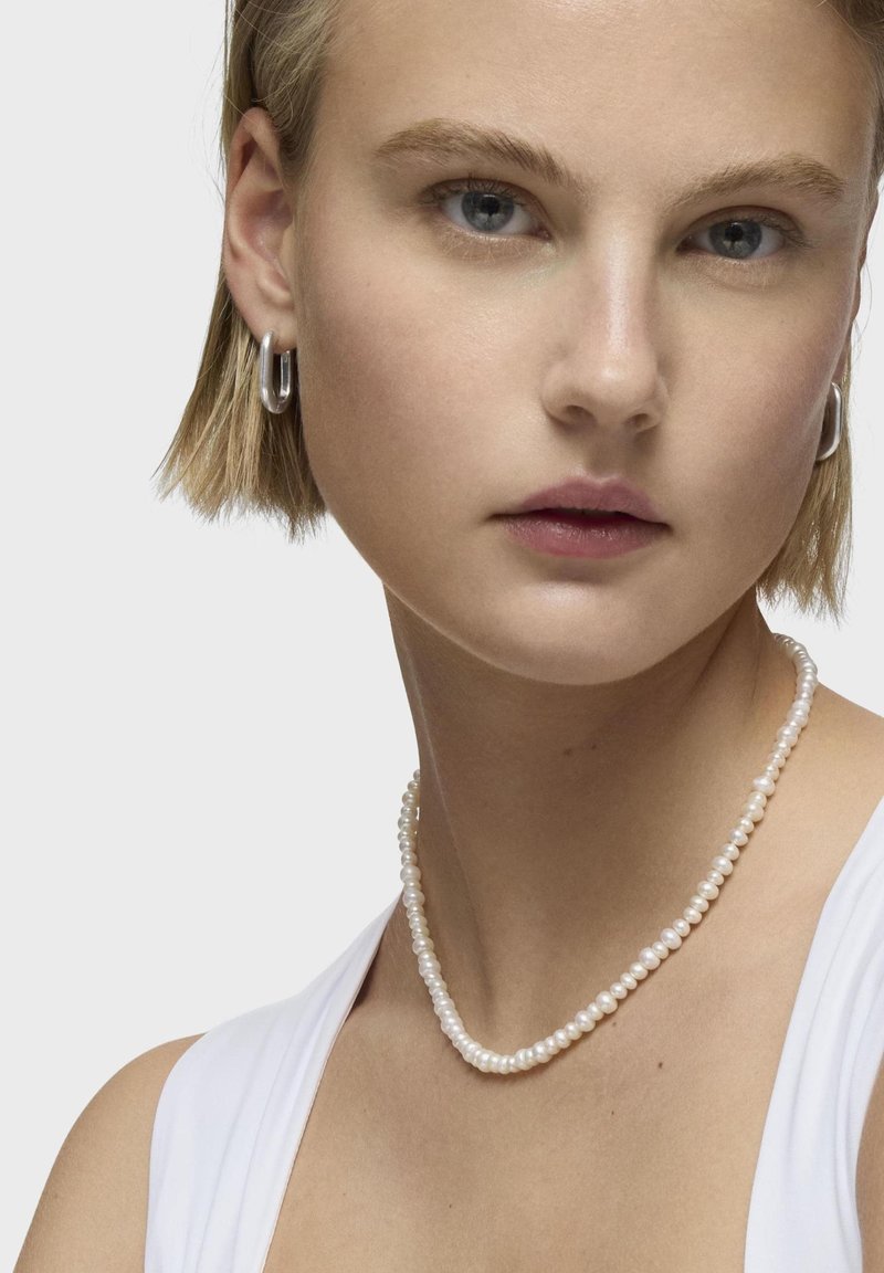 Collana di perle con piccole perle bianche rotonde, superficie liscia, indossata con una semplice maglietta bianca. Orecchini a cerchio in argento completano il look.