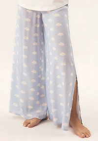 Jasnoniebieskie spodnie o szerokich nogawkach z przezroczystego materiału, z powtarzającym się białym wzorem chmur i bocznymi rozcięciami, z odkrytymi stopami na dole.