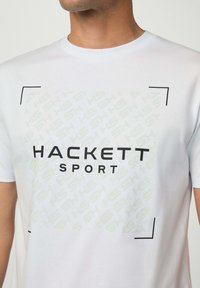 Bílý bavlněný tričko s grafickým potiskem s texty "HACKETT" a "SPORT" v černé barvě, obdélníkovými akcenty a decentním vzorovaným pozadím.