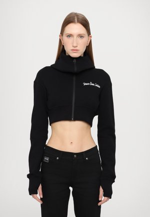 Schwarzer cropped Sweatshirt mit hohem Kragen, vorderem Reißverschluss und gesticktem Logo. Lange Ärmel mit Rippbündchen, kombiniert mit schwarzen Jeans.