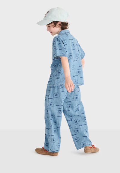 Enfant portant une tenue en denim bleu clair avec des imprimés de dessins animés, une casquette beige et des chaussures marron, marchant de côté sur un fond uni.