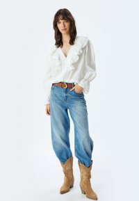 Witte blouse met ruches kraag en knoopdetails, gecombineerd met blauwe high-waisted jeans en tan cowboylaarzen met decoratieve stiksels.