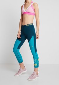 Kvinna i rosa sport-bh, turkosa och marinblå Nike Pro leggings med gula detaljer, samt ljusrosa träningsskor, står framför en grå bakgrund.