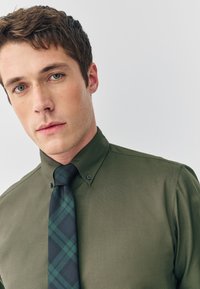 Camisa verde abotonada con textura lisa, combinada con una corbata de cuadros en tonos negro y verde. El cuello es abotonado con costuras visibles.