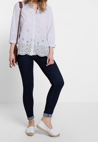 Weiße Bluse mit langen Ärmeln und floralem Lochmuster am Saum, kombiniert mit eng geschnittenen Jeans aus dunkelblauem Denim und weißen Espadrilles.