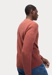 Suéter de punto en color óxido con un patrón texturizado, mangas largas y un cuello redondo, combinado con jeans azules.