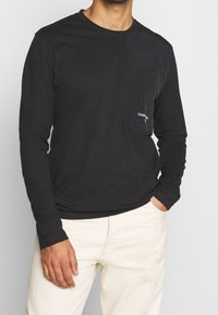 Černé tričko s dlouhým rukávem, s kapsou na levé straně hrudi a detailem loga. Vyrobeno z měkkého materiálu, jednoduchý design a přiléhavý střih.