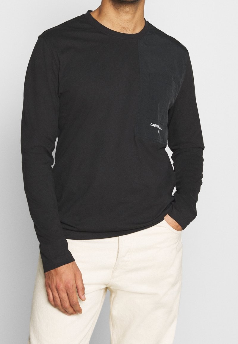 Černé tričko s dlouhým rukávem, s kapsou na levé straně hrudi a detailem loga. Vyrobeno z měkkého materiálu, jednoduchý design a přiléhavý střih.