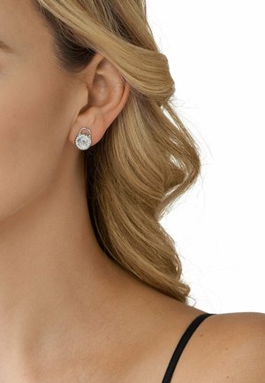 Silberne Ohrringe mit einem runden, klaren Edelstein und einem dekorativen Halo, auf einem Modell mit welligem, blonden Haar gezeigt.