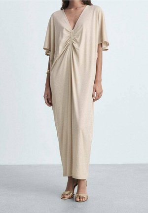 Un mannequin porte une robe beige ample jusqu’aux chevilles, avec un col en V, un devant froncé, des manches courtes et des sandales dorées.