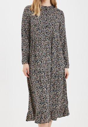 Kvinde iført en langærmet, sort midi-kjole med små pink og hvide blomsterprint, stående mod en ensfarvet hvid baggrund.