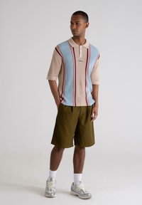 Knitted polo shirt in light blue and beige with maroon stripes, three-button placket, paired with olive green shorts and gray sneakers.