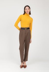 Gelber Rollkragenpullover und braune, hoch taillierte Hose mit einem schwarzen Gürtel. Die Schuhe sind braune, spitze Ballerinas. Modell steht vor einem weißen Hintergrund.